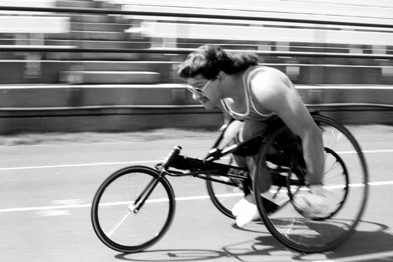 Frank Epperson speeds down the Michael A. Carroll Track in his wheelchair as part of the 1992 Paralympic Games. Black and white photo.